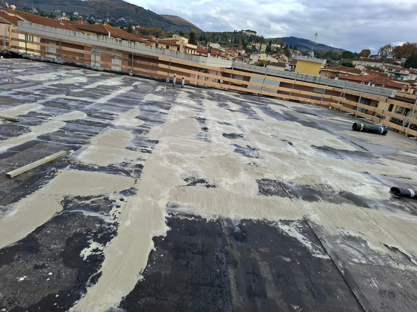 Un lotto di asfalto vuoto con riparazioni bianche a chiazze, accanto a edifici bassi e colline sotto un cielo nuvoloso.