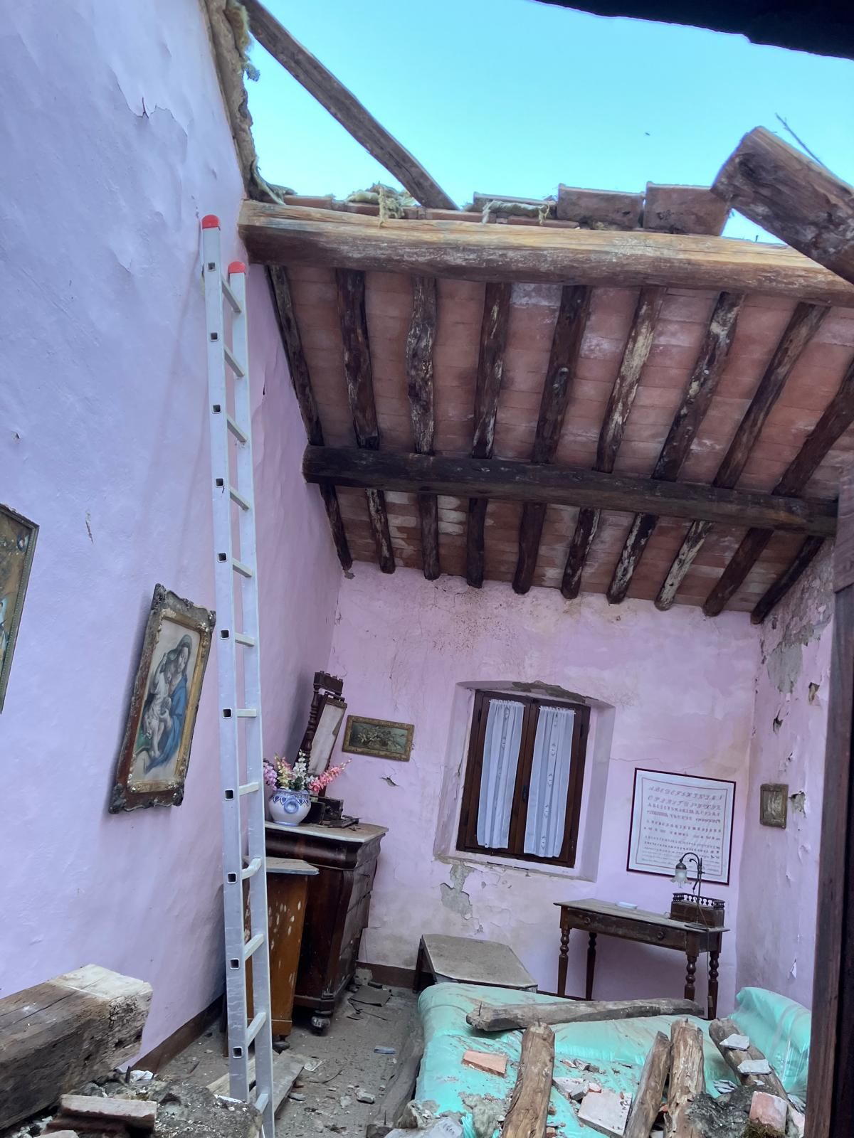 Stanza rosa parzialmente crollata con travi di legno a vista sul soffitto, scala e detriti sparsi sul pavimento.