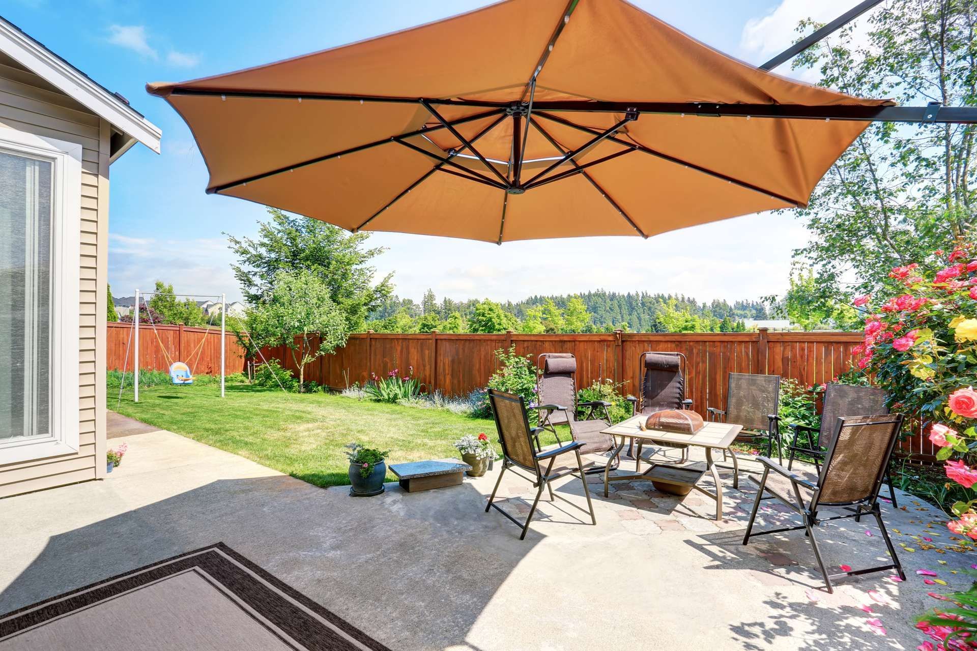 A tan umbrella shades a patio with outdoor chairs and a table in a residential backyard with a wood fence and green lawn.