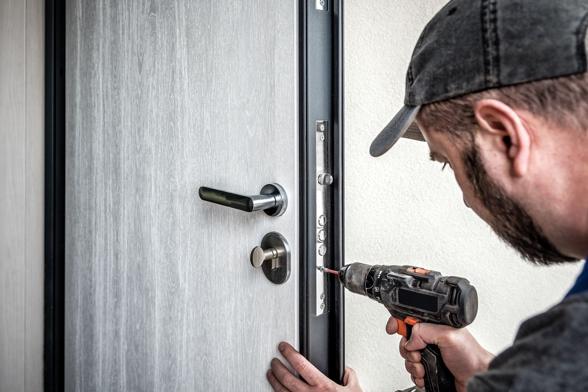 A person wearing a cap uses a power drill to install or repair a lock on a light-colored wood door.