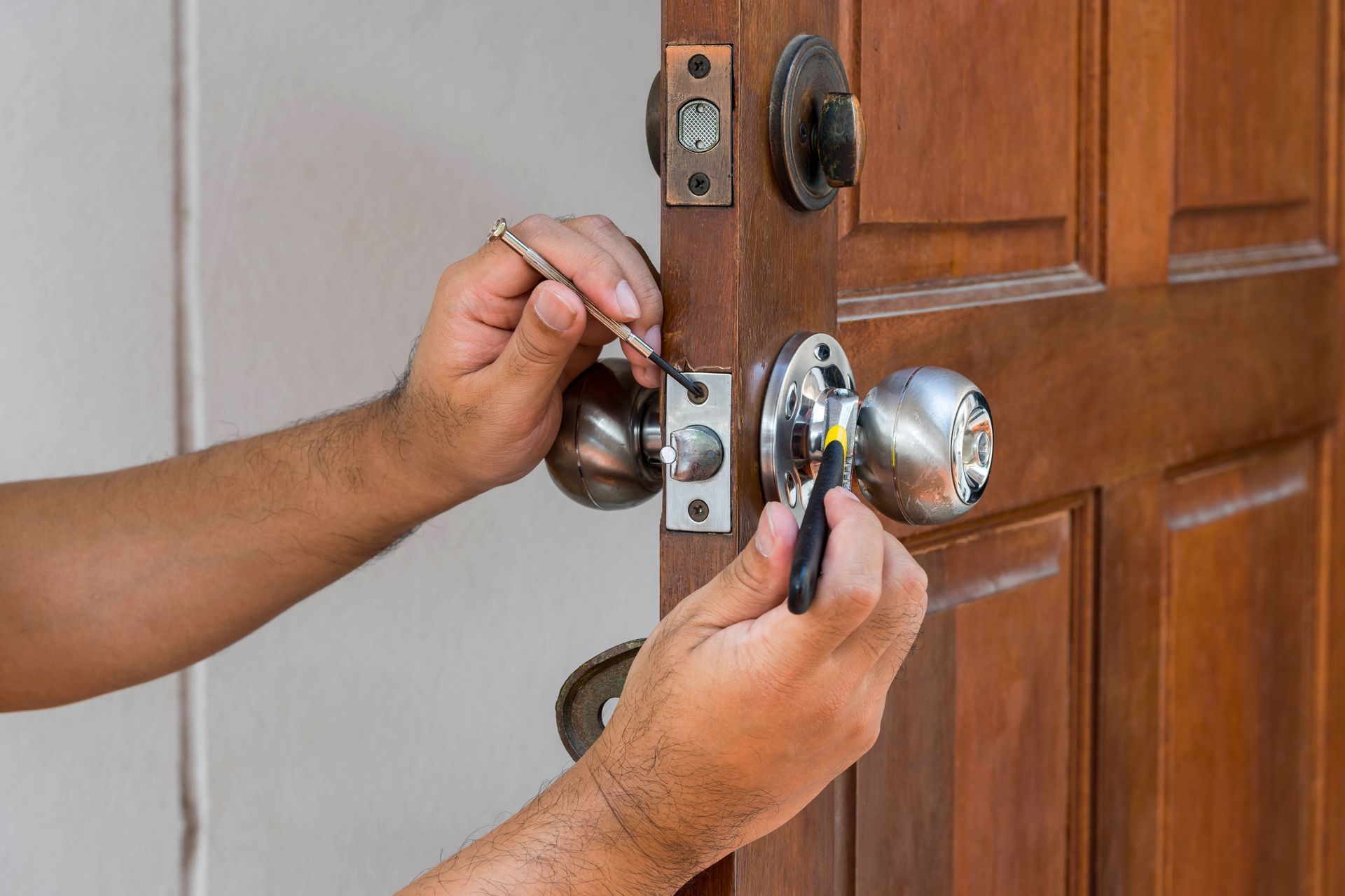 Close-up of hands using a screwdriver and a tool to repair or install a metal doorknob on a wooden door.