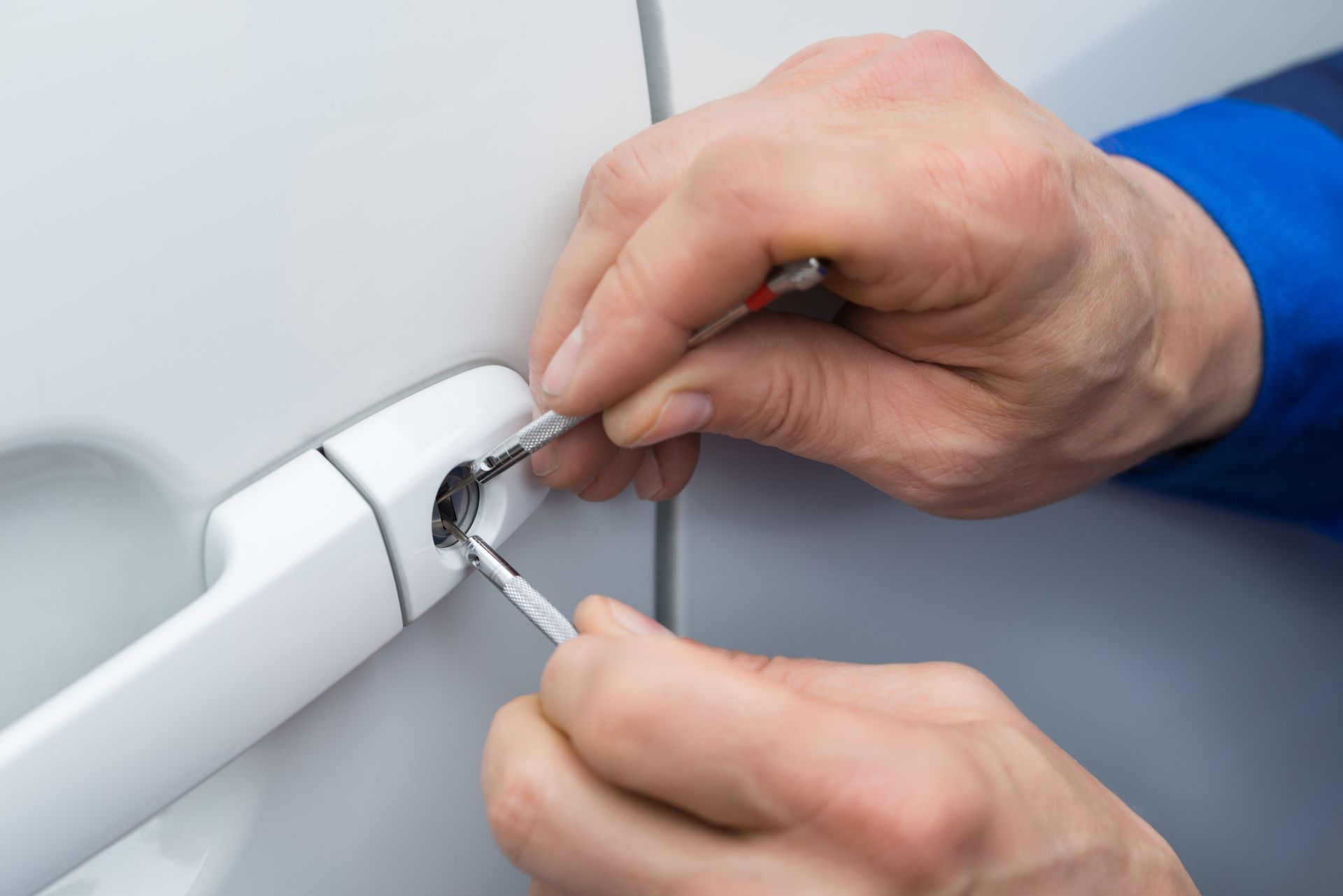 A pair of hands using metal tools to pick the lock on a white car door handle.