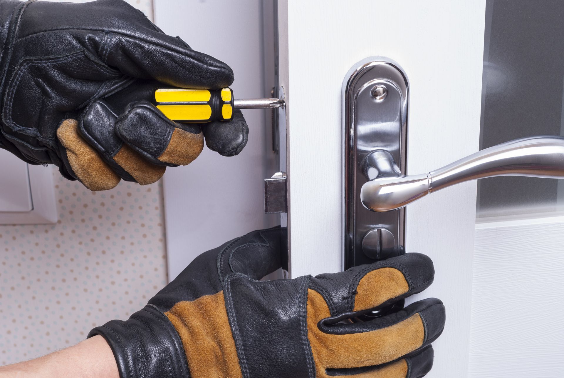 Hands wearing work gloves using a yellow-handled screwdriver to install or adjust a metal door handle on a white door.