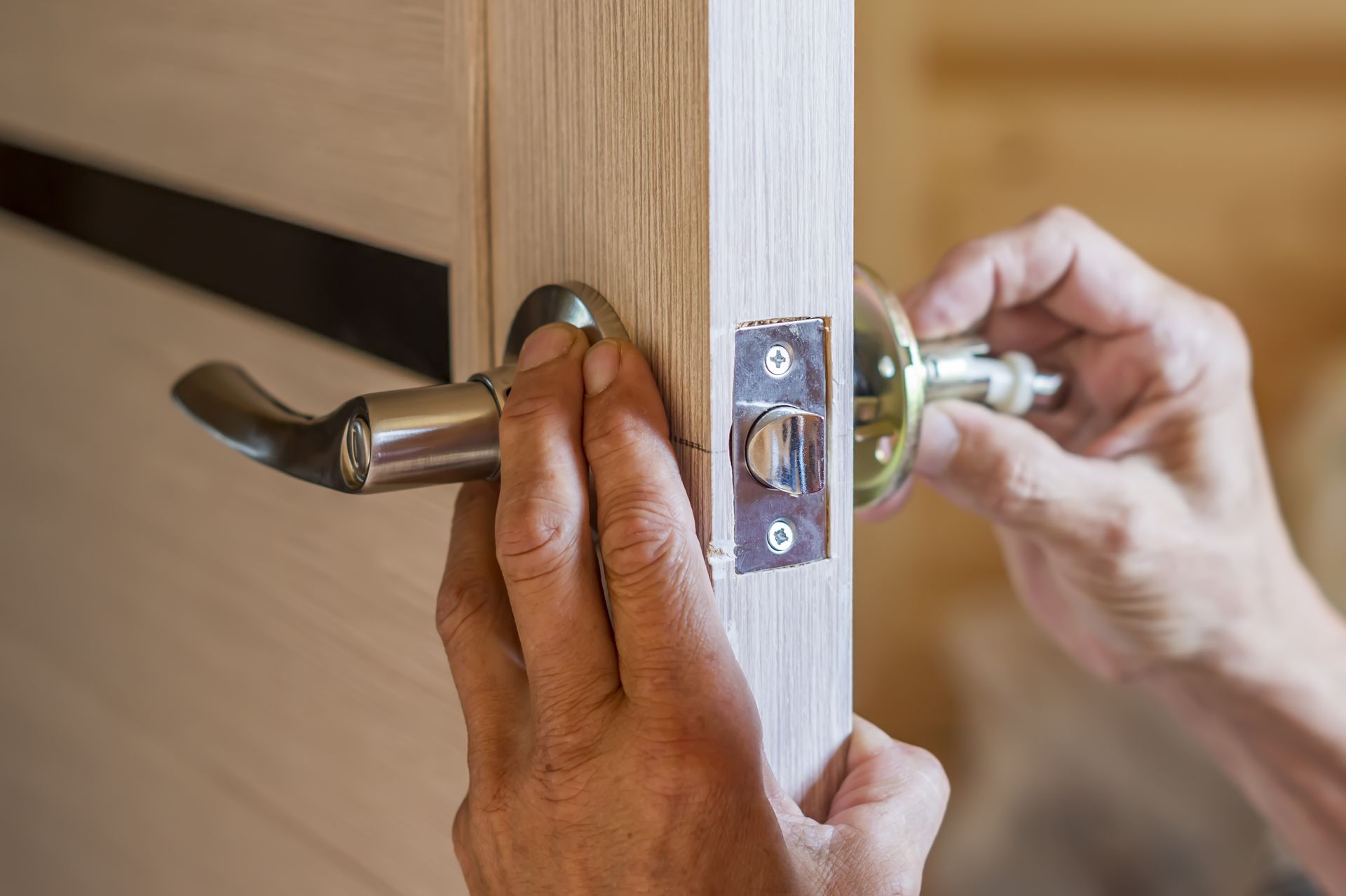 Hands installing a silver metal door handle and latch mechanism onto a light wood door.