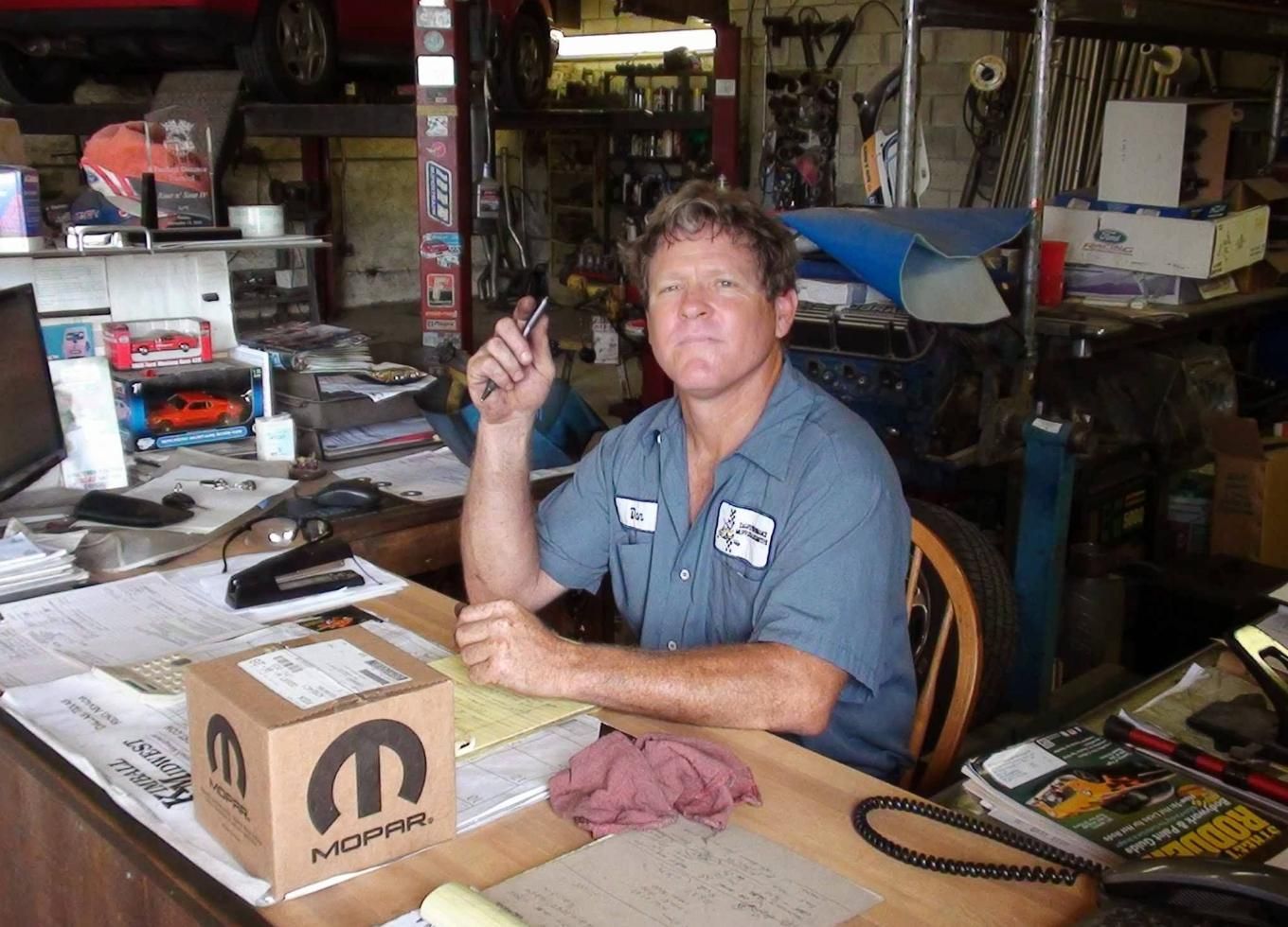 Man sitting at a cluttered workbench holding a tool in a busy garage workshop