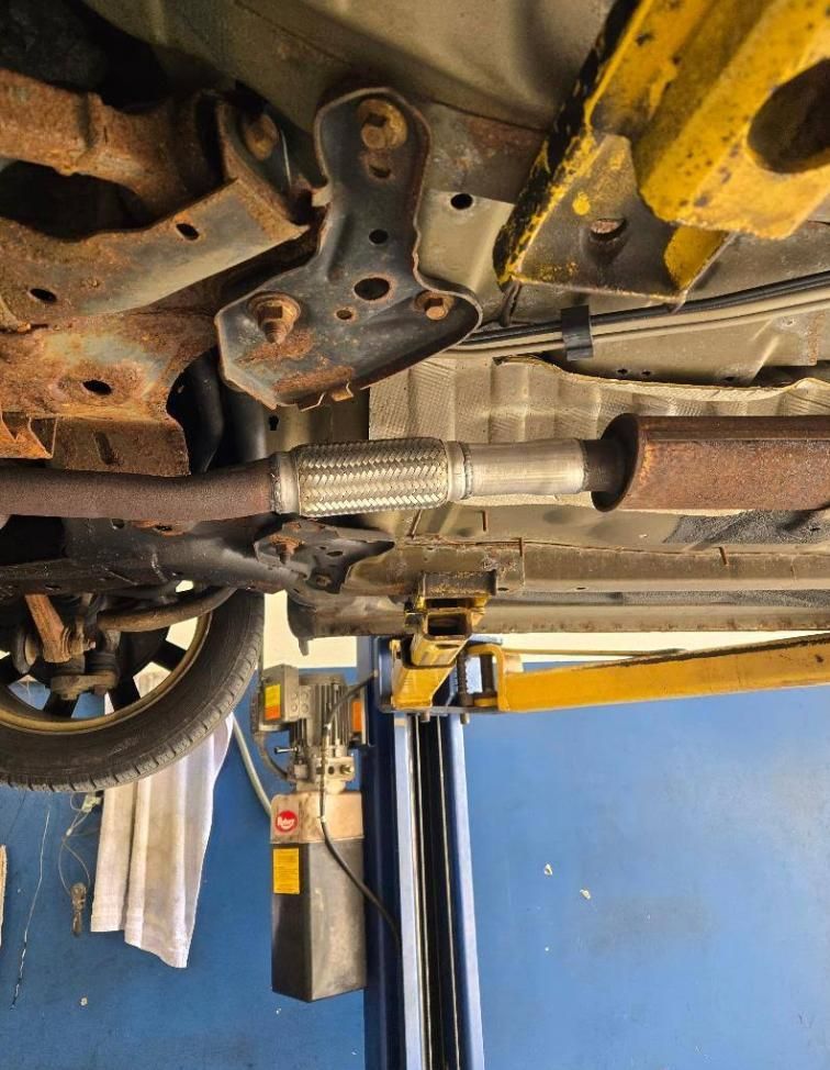Underside of a car on a lift, showing rusted exhaust piping and a mechanic’s shop background