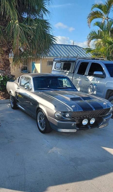 Classic silver muscle car with black racing stripes parked in a driveway beside an SUV, palm trees behind it