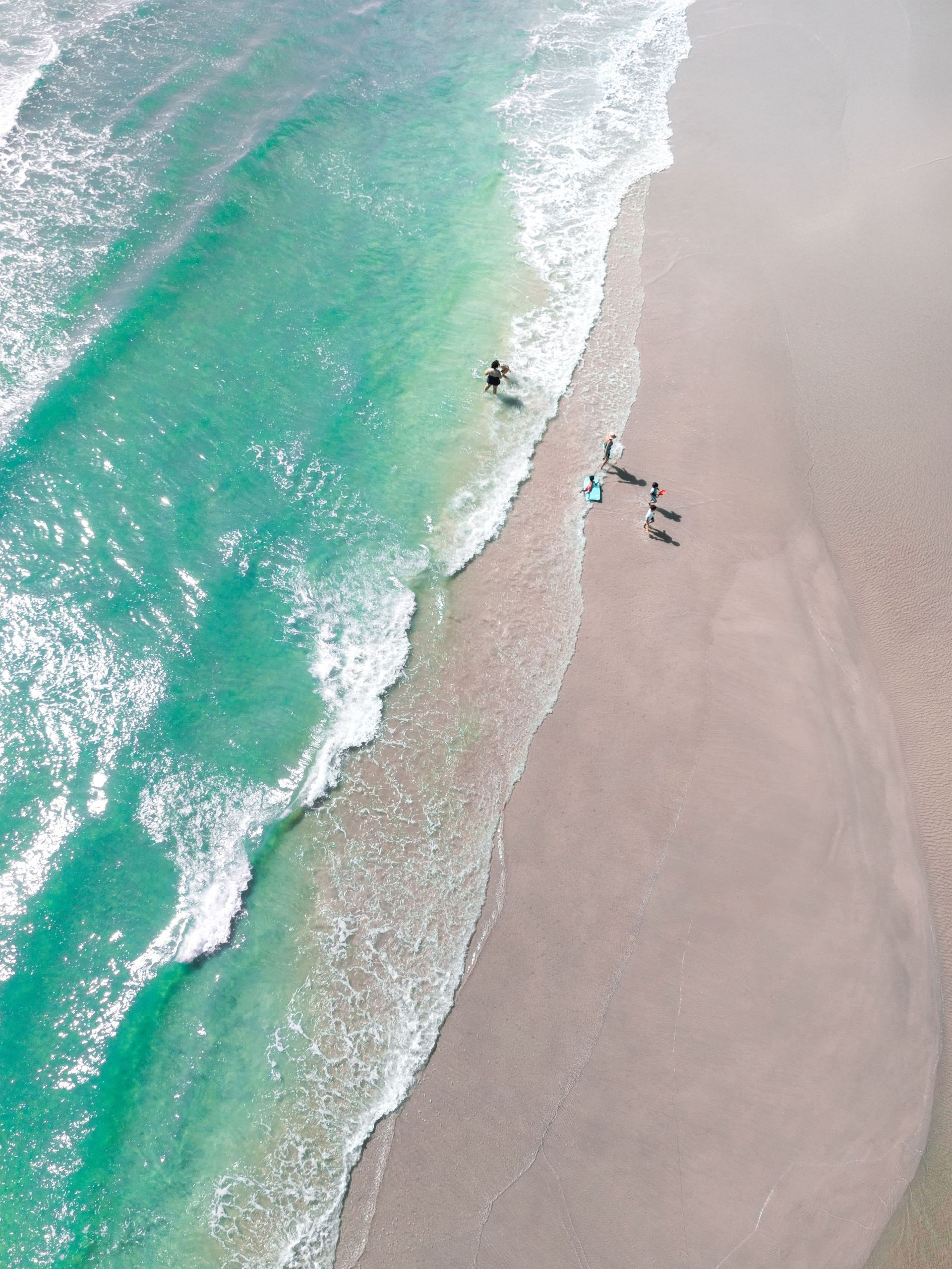 Aerial view of turquoise waves washing onto a pale sandy beach with a few people near the shoreline.