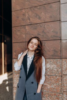 Woman in a gray vest leaning against a stone wall, smiling softly outdoors.