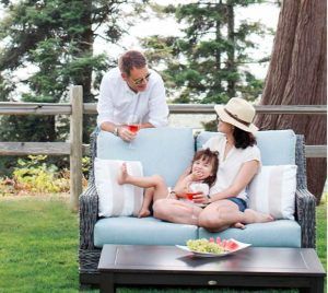 Family relaxing on an outdoor couch in a garden, with drinks and a plate of fruit on the table