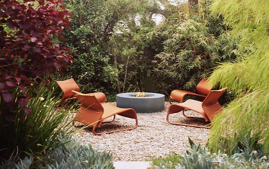 Modern patio seating with orange chairs around a round fire pit, surrounded by lush garden greenery.