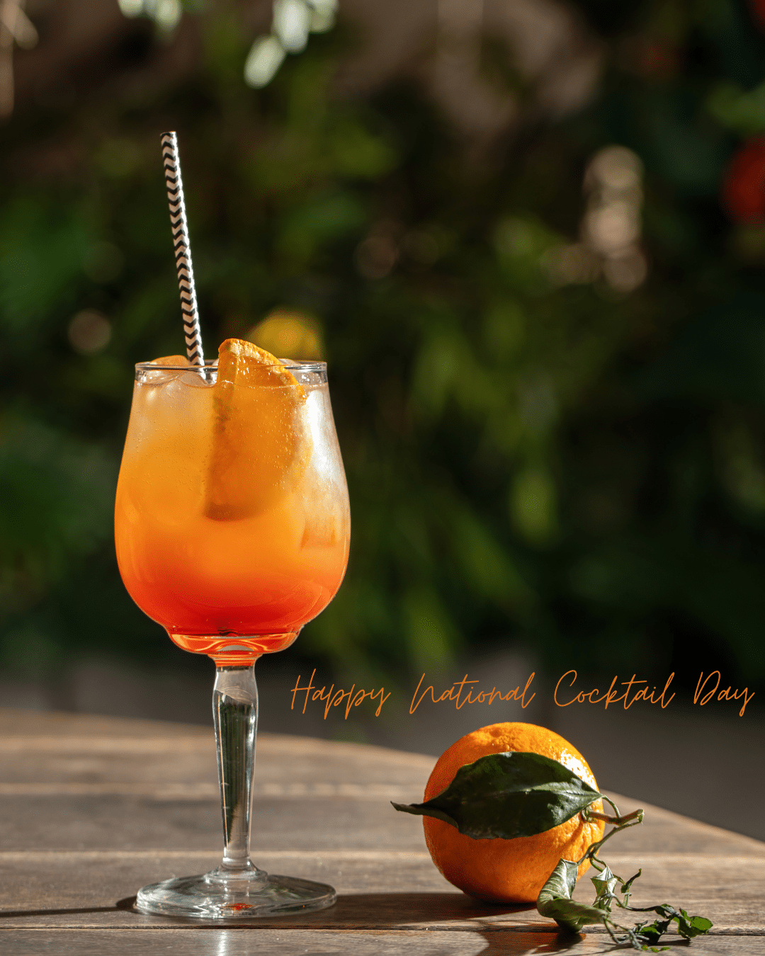 Orange cocktail in a stemmed glass with a striped straw beside a cut orange on a wooden table.