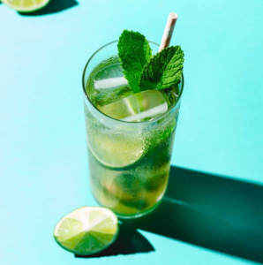 Mint lime drink in a glass with ice on a turquoise surface, with a lime wedge nearby.