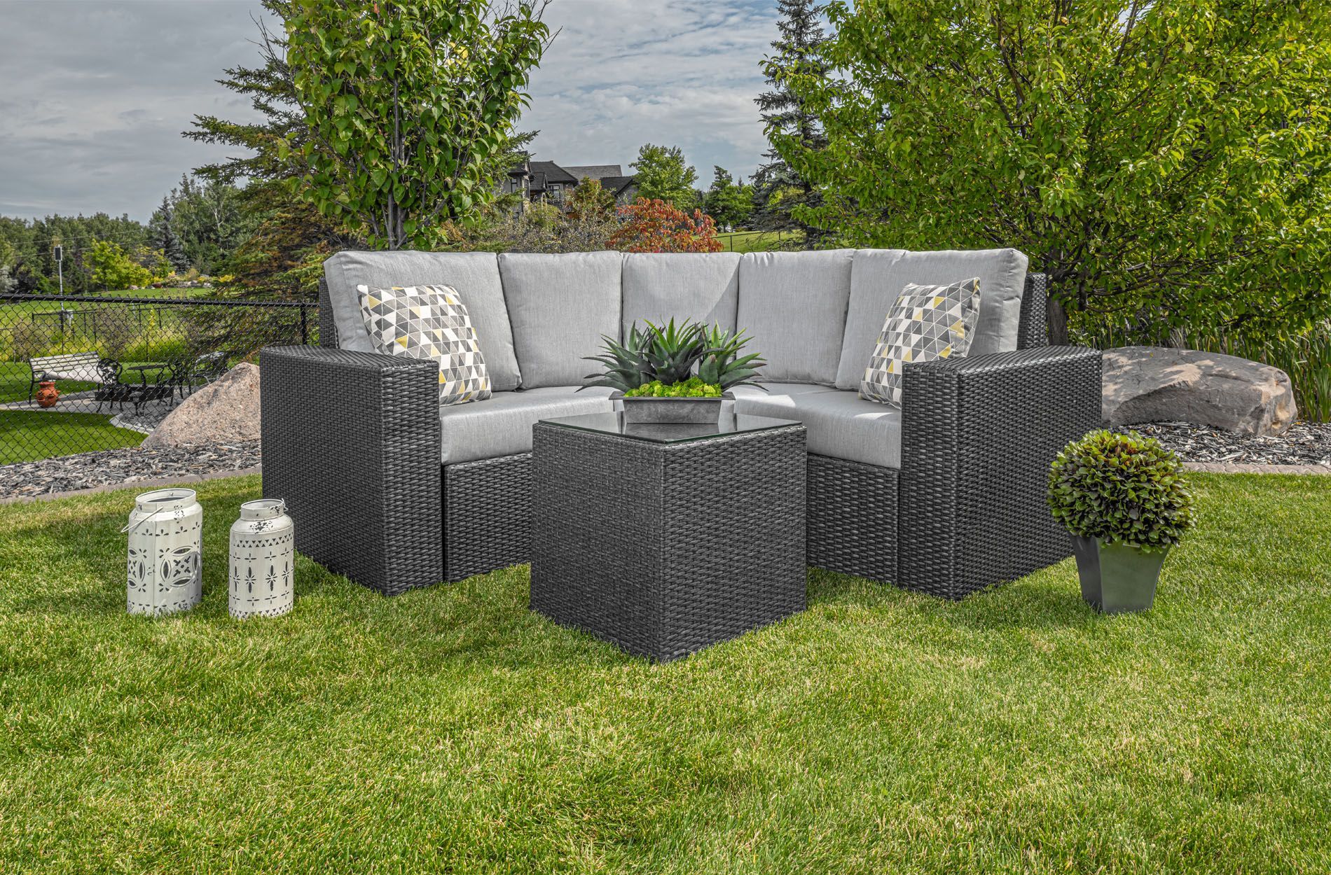 Outdoor patio set with gray wicker sofas, black table, and potted plants on a grassy lawn.