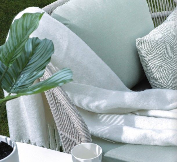 A white wicker chair with pale green pillows, a light blanket, and a leafy plant in a soft, cozy corner.
