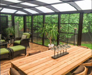 Sunroom with wicker chairs, green cushions, potted plants, and a wooden table under a glass roof.
