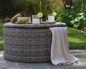 Round wicker outdoor table set with drinks, candles, and a draped white throw on a patio lawn.