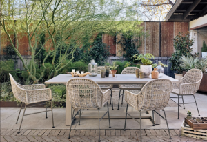 Outdoor patio dining area with wicker chairs, a long table, and lush garden greenery under a pergola.