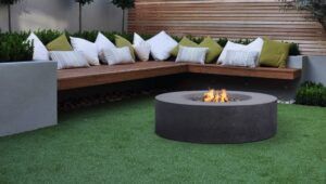 Modern patio with curved bench seating, white and green cushions, and a round fire pit on artificial grass