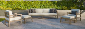 Outdoor patio seating set with beige sofas and armchairs on a stone terrace beside a green hedge