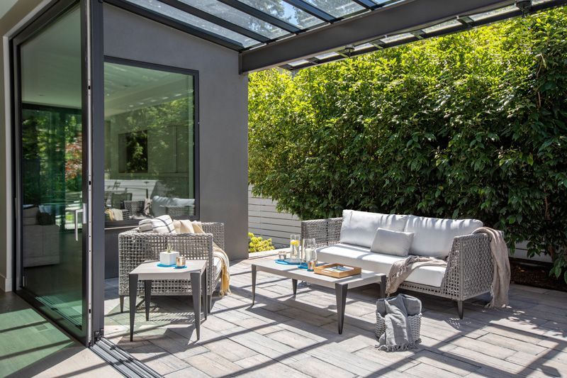 Modern patio with gray sofa, coffee table, and glass roof beside a lush green hedge