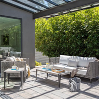 Patio lounge with wicker sofa, coffee table, and chairs under a glass canopy beside a green hedge.