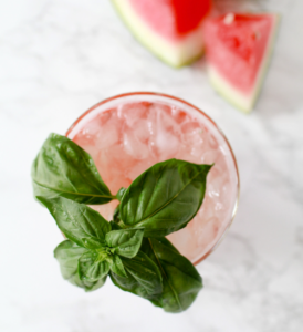 Pink cocktail with mint leaves and ice, garnished on a light surface with watermelon slices in the background