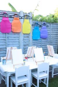 Outdoor dining table with white chairs under colorful hanging lanterns beside a gray fence