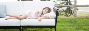 Person sleeping on a white couch outdoors on a sunny lawn, with a pillow and blanket.