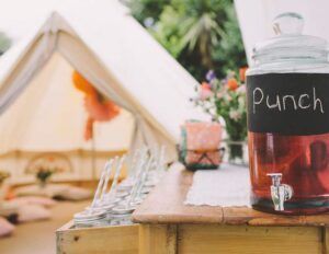 Outdoor picnic table with a glass of punch, utensils, and a blurred white tent in the background