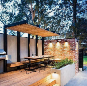 Modern outdoor patio with wooden pergola, dining table, brick wall lights, and built-in planter seating.