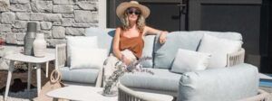 Woman in a straw hat relaxing on a light blue sofa on a sunlit patio with stone walls.