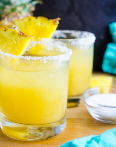 Two yellow cocktails with pineapple garnish and salted rims on a wooden table
