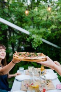 Friends sharing a pizza outdoors at a garden table with drinks and plates