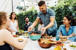 Friends sharing food at an outdoor picnic table, with one person serving from bowls and dishes.