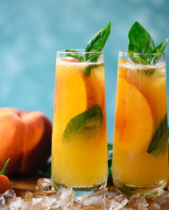 Two tall peach drinks with mint leaves and ice on a wooden surface, with peaches in the background