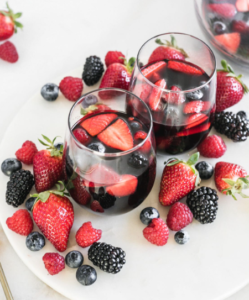 Two glasses of red berry drink surrounded by strawberries, blueberries, and blackberries on a white table.