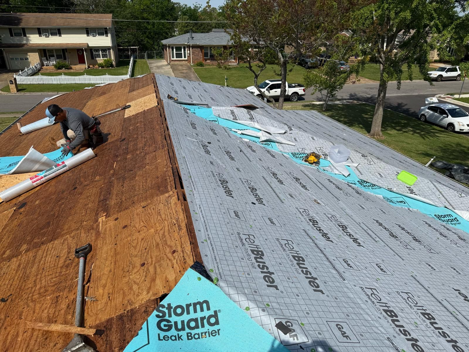 Roofer installing roofing materials, with blue underlayment and plywood, on a house in a residential area.