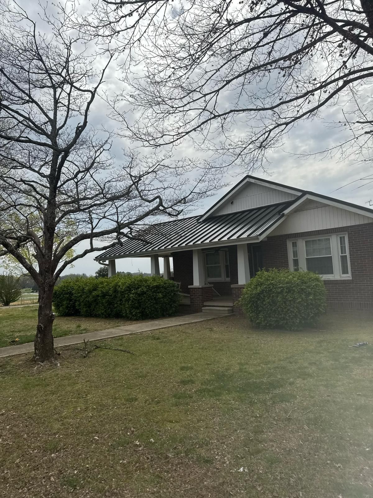 Brick house with dark metal roof, porch, and two shrubs, under a cloudy sky.