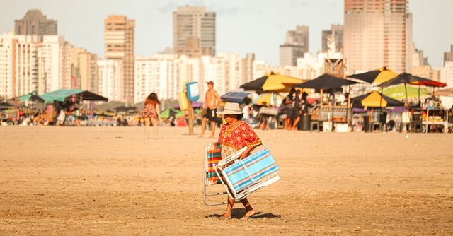 Una persona empuja un carrito a rayas por una playa de arena con sombrillas y edificios de la ciudad al fondo.