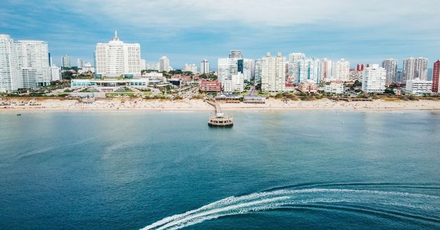 Horizonte de una ciudad costera con edificios blancos, un pequeño barco en aguas azules tranquilas y una estela.