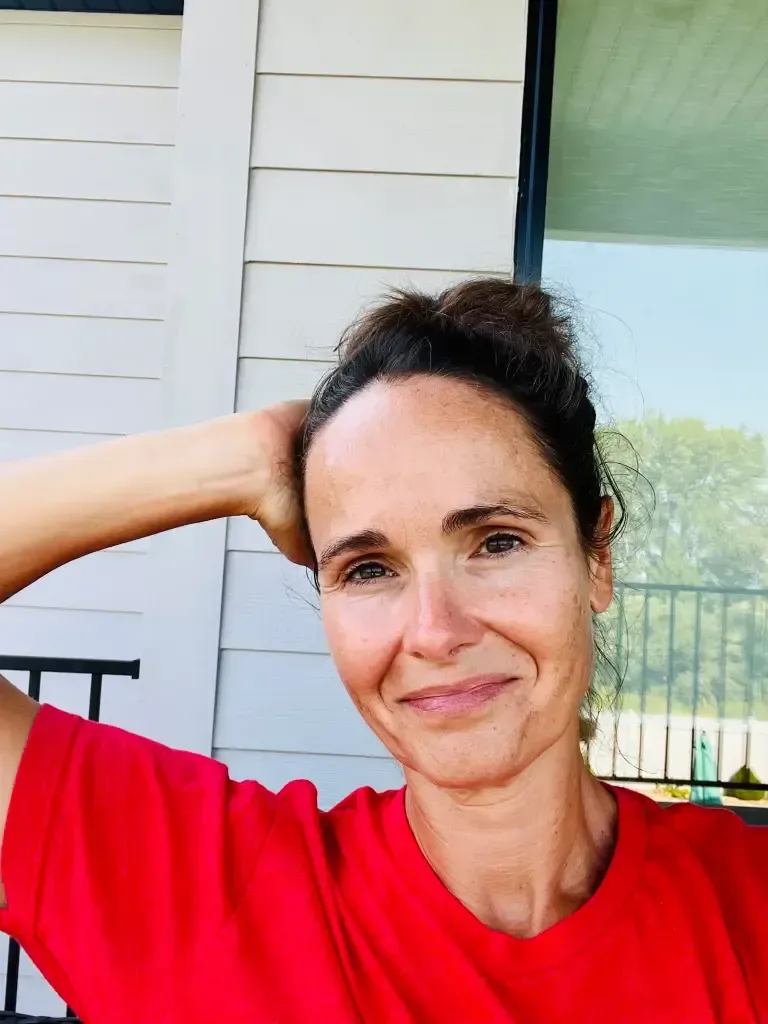 A woman in a red shirt is smiling with her hand on her head.