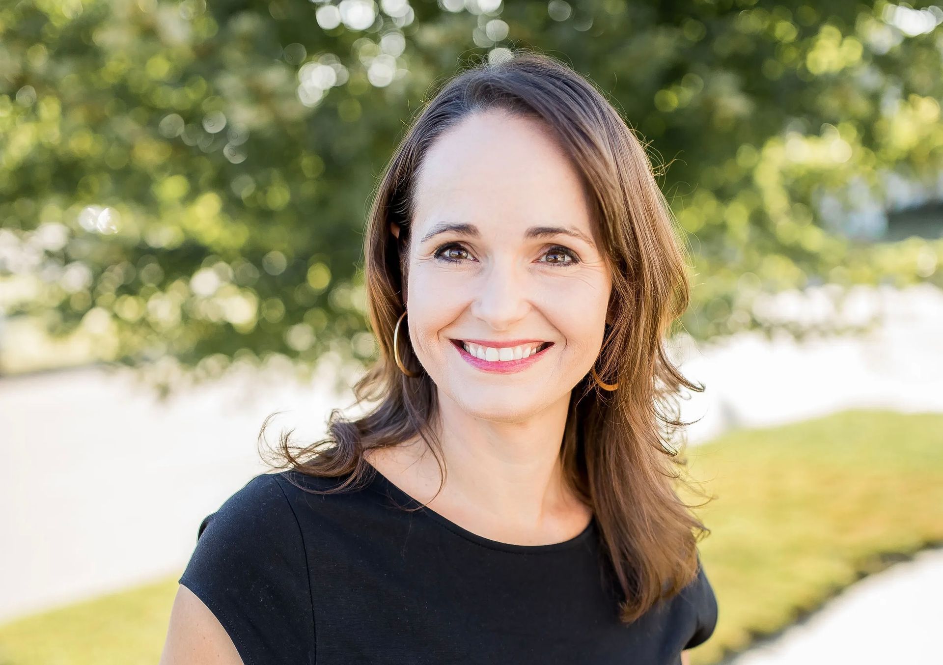 A woman in a black shirt is smiling for the camera while standing in front of a tree.