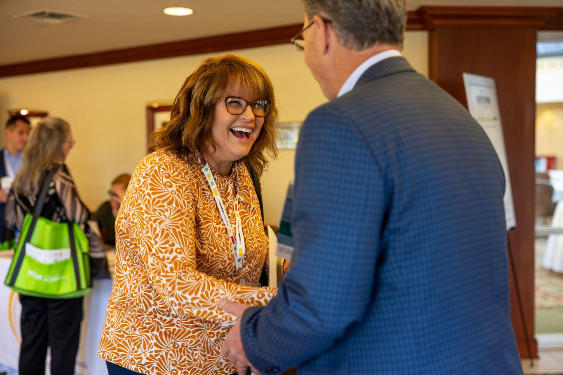 A smiling person wearing a patterned orange top shakes hands with someone in a blue suit at an indoor event.