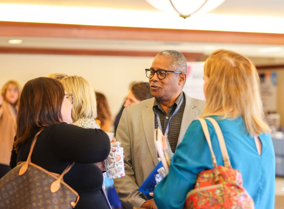 A group of people in business-casual attire talk and smile in a bright, indoor hallway at a professional conference.
