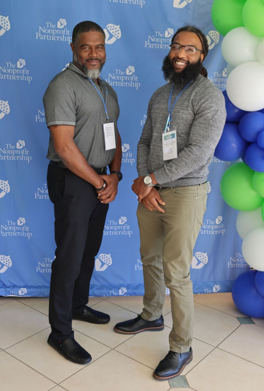 Two individuals in casual business attire pose in front of a blue backdrop with event branding and green and blue balloons.