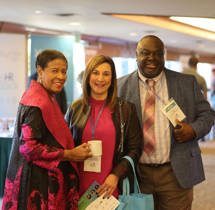 Three people pose for a photo at an indoor professional event, smiling and holding drinks and a name tag.