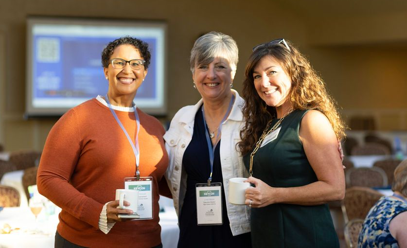 Three people smiling and posing at a conference, holding drinks and wearing name badges.