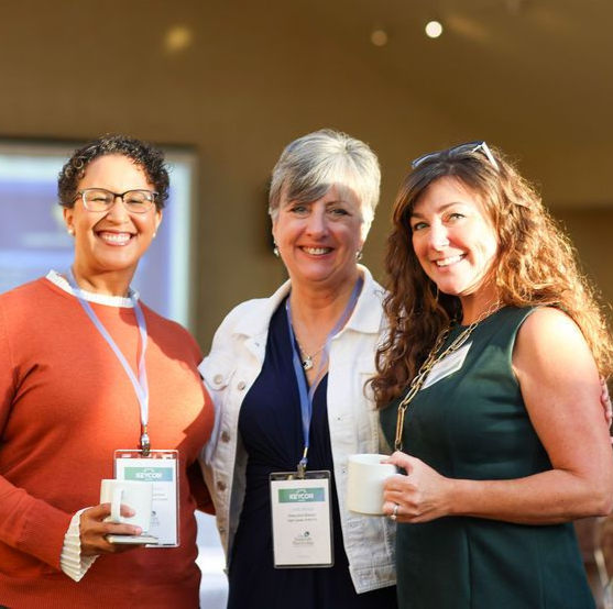 Three smiling individuals stand together at an indoor event, wearing conference badges and holding a white mug.