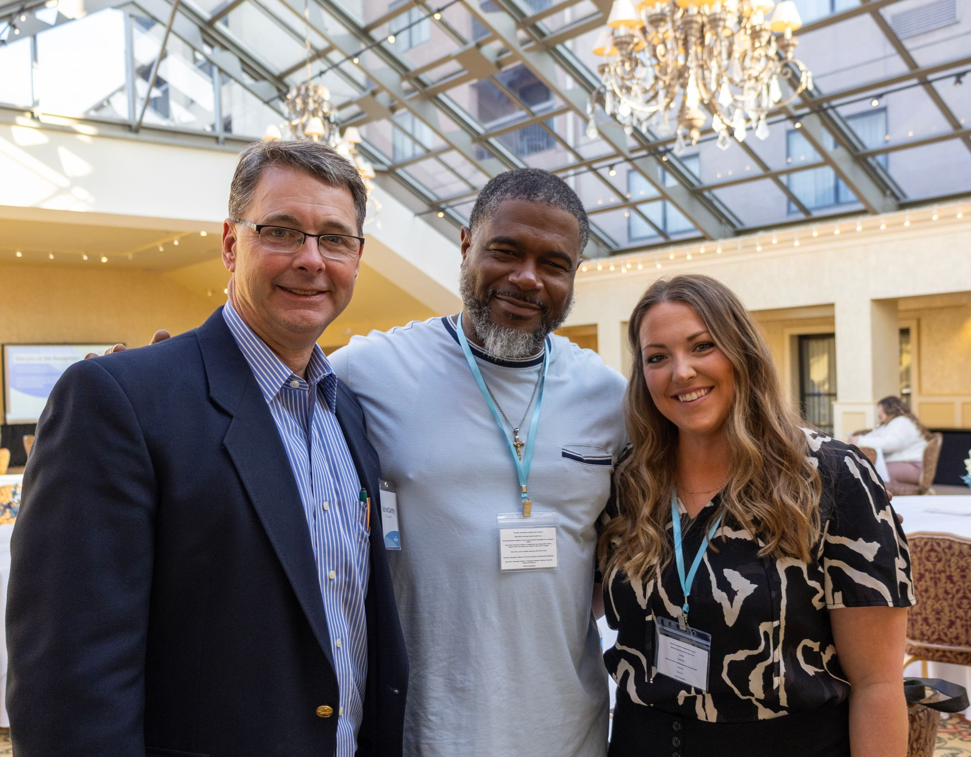 Three people pose for a photo in a bright, indoor atrium with a large glass ceiling and a hanging chandelier.